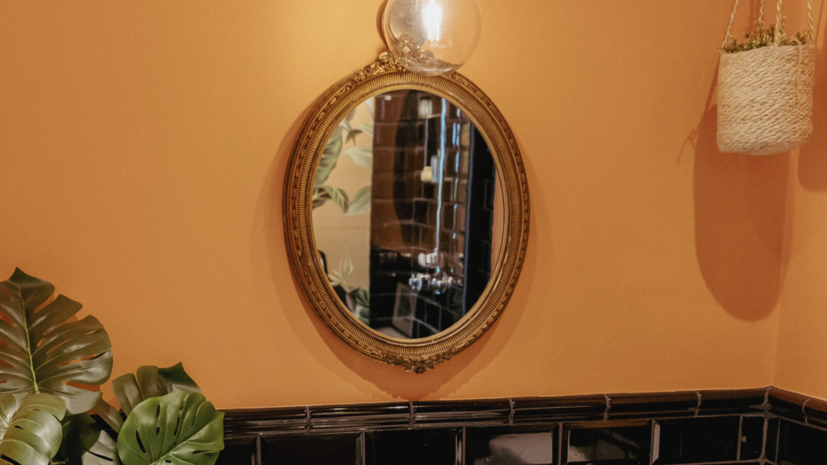 A gold framed mirror against a bright orange wall.