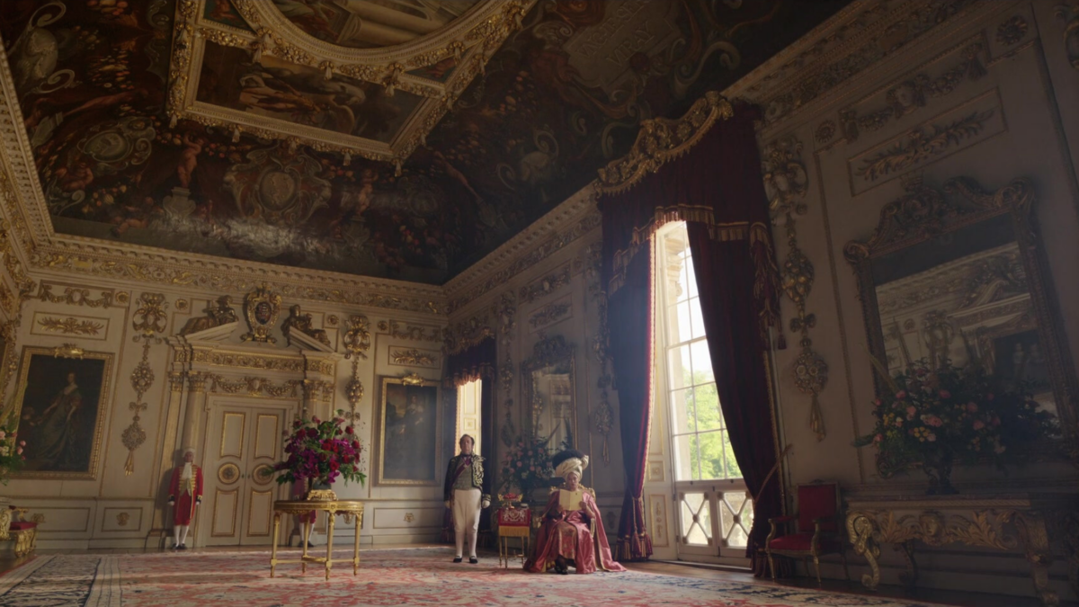 A wideshot of a room in Bridgerton with a painted ceiling and gold trimming on the ceiling and the walls. Queen Charlotte sits in the centre.