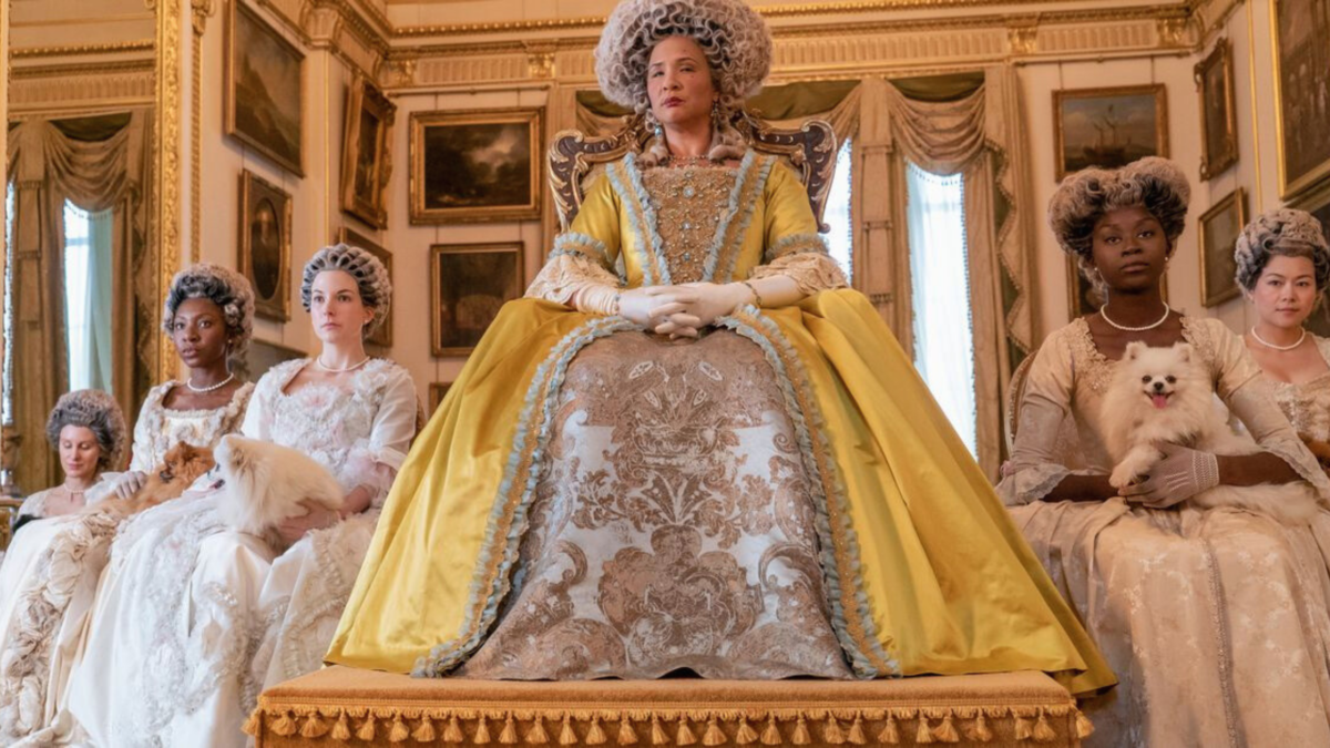 Queen Charlotte sitting in the middle of the frame with three women to one side of her and two to the other side. In the background, there are paintings on the wall.